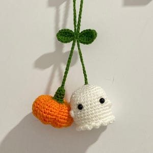 May include: Crocheted Halloween decorations. The image shows a small orange pumpkin and a white ghost with black eyes. They are connected by green yarn and a green leaf. Ideal for Halloween.