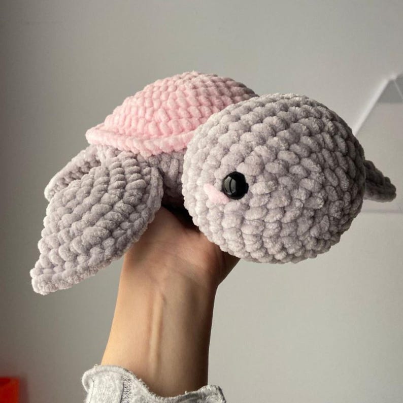 May include: A handmade crochet turtle plushie. The turtle has a gray body, flippers, and head, with a pink shell. The eyes are black, and the plushie is made of soft, textured yarn. A cute and cuddly stuffed animal.