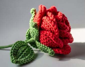 May include: A handcrafted crochet rose featuring vibrant red yarn petals arranged in a layered bloom. The flower includes a green stem, textured green sepals, and a single detailed green leaf attached to the base of the stem.