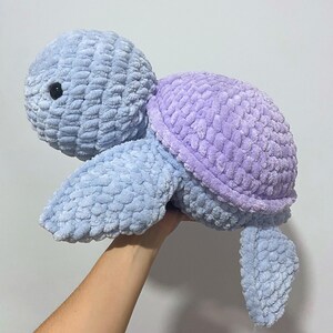 May include: A plush, crocheted turtle in shades of blue and lavender. The turtle has a textured, soft appearance with a rounded head, shell, and flipper-like limbs. The eyes are small and black. The turtle is held up by a hand.