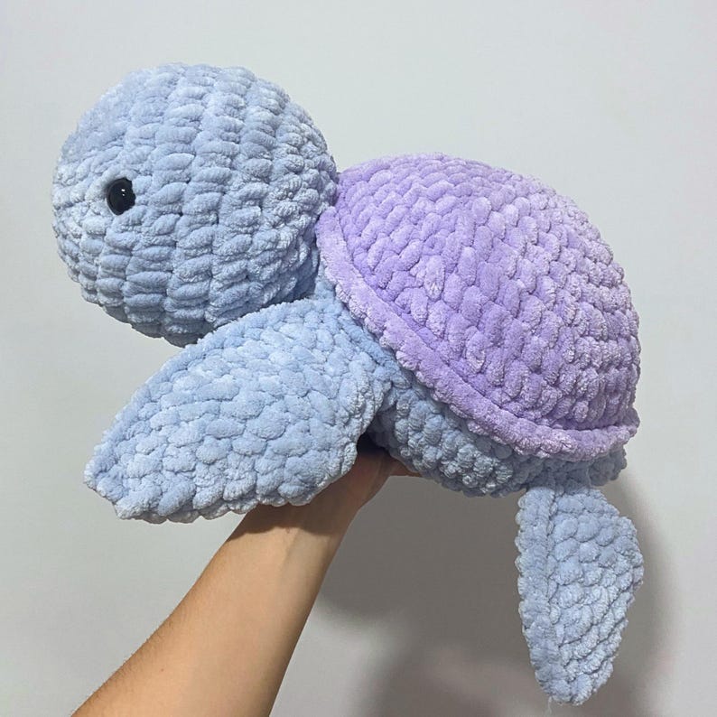 May include: A plush, crocheted turtle in shades of blue and lavender. The turtle has a textured, soft appearance with a rounded head, shell, and flipper-like limbs. The eyes are small and black. The turtle is held up by a hand.
