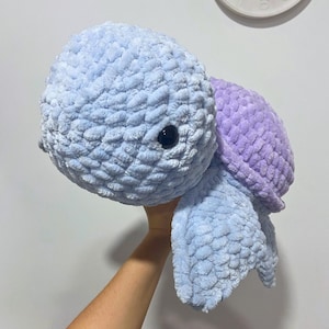 May include: A handmade plush turtle in shades of blue and purple. The turtle has a textured, soft appearance, with a light blue head and limbs, and a purple shell. It features a pair of black, round eyes. The plush toy is designed to be cuddly.