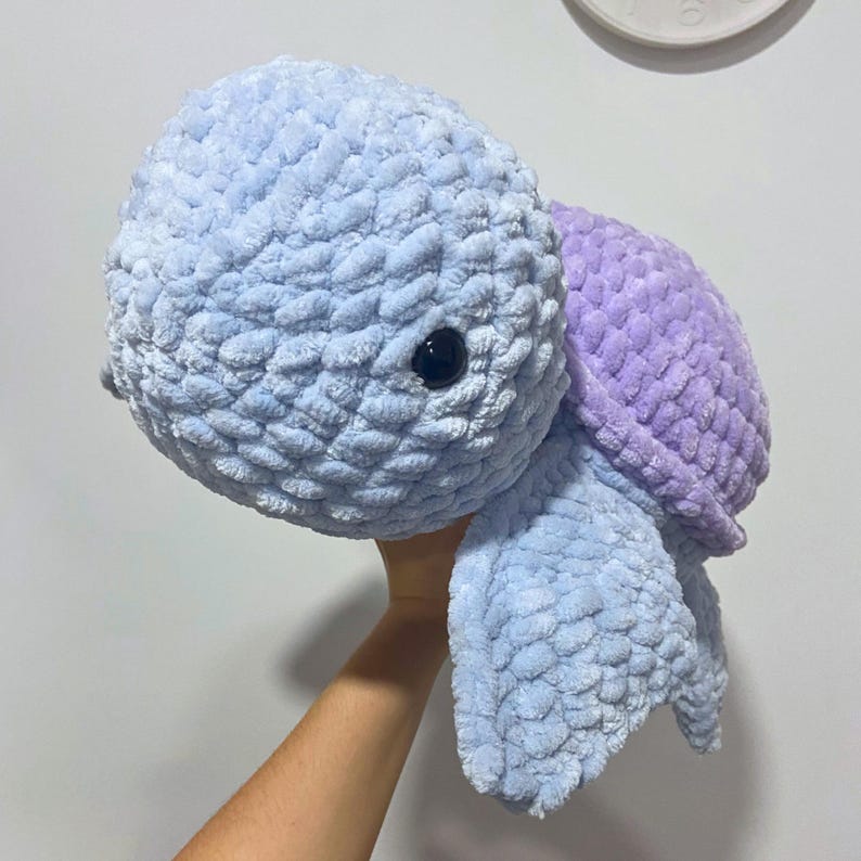 May include: A handmade plush turtle in shades of blue and purple. The turtle has a textured, soft appearance, with a light blue head and limbs, and a purple shell. It features a pair of black, round eyes. The plush toy is designed to be cuddly.