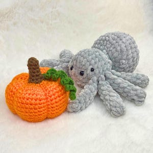 May include: A crocheted Halloween-themed set featuring a plush gray spider and an orange pumpkin with a brown stem and green vine. The spider has eight legs and small black eyes. The pumpkin is round and textured.