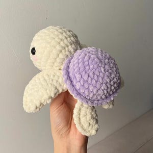 May include: A handmade crochet turtle plushie in cream and lavender. The turtle has a cream-colored head, flippers, and body, with a lavender shell. The turtle has black eyes and a pink blush on its cheeks. The plushie is soft and textured.