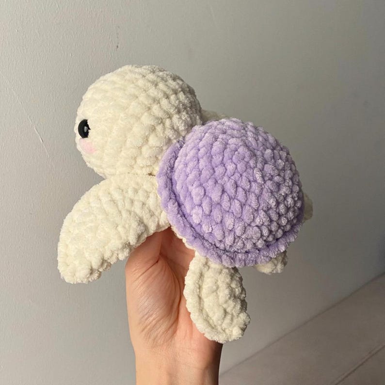 May include: A handmade crochet turtle plushie in cream and lavender. The turtle has a cream-colored head, flippers, and body, with a lavender shell. The turtle has black eyes and a pink blush on its cheeks. The plushie is soft and textured.