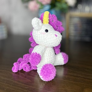 May include: A white crocheted unicorn with a purple and yellow mane and tail. The unicorn is sitting on a wooden surface.