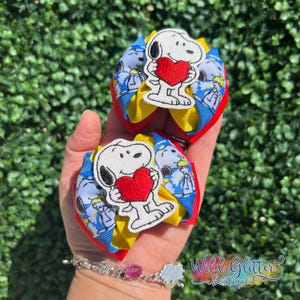 May include: Two Snoopy hair bows with red, yellow, and blue ribbons. Each bow features a Snoopy applique holding a red heart. The bows are held in a hand against a green background.