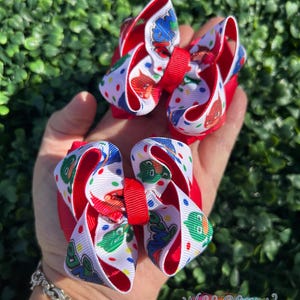 May include: Three red and white hair bows with a cartoon character print. The bows have a red base and center, with a white ribbon featuring colorful character designs and polka dots. The bows are held in a hand, with a bracelet visible.