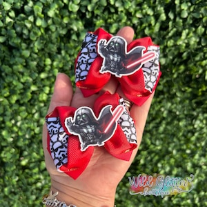 May include: Two red hair bows with a Star Wars theme. Each bow features a Darth Vader sticker with a red lightsaber and a black and white patterned accent. The bows are set against a green background.