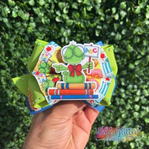 May include: A hair bow featuring a green cartoon character wearing glasses and a red bow tie, sitting on a stack of three books. The character holds a sign with the text ABC. The ribbon has a pattern of books, apples, and pencils on a white background.