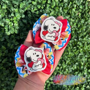 May include: Two handmade hair bows featuring Snoopy holding a heart. The bows have red, white, and blue chevron and character-themed ribbons. The Snoopy applique is white with black ears and a pink bow.