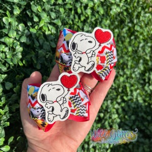 May include: Two handmade hair bows featuring Snoopy holding a red heart balloon. The bows have a red base with a chevron pattern in white, black, and yellow. The Snoopy applique is white with black detailing.