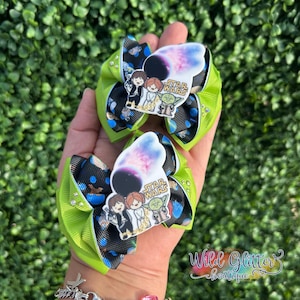 May include: Two Star Wars themed hair bows. Each bow features a green and black patterned base with a Star Wars graphic. The graphic includes characters and the Star Wars logo. The bows are held in a hand against a green background.