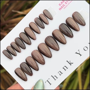 May include: A collection of press-on nails in different sizes, displayed on a white card. The nails have a shimmering, metallic brown finish. The card features the text "Thank You" and "Kar Nail Studio Press on Nails".
