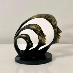 May include: Abstract sculpture of three faces, one inside the other, with a black and gold finish. The sculpture is made of resin and is a modern and stylish home decor piece.