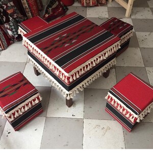 May include: A set of four red and black patterned square ottomans with white fringe. The ottomans are arranged in a square shape with a larger ottoman in the center.