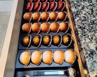 May include: A drawer filled with a variety of coffee pods. The pods are arranged in rows and are in different colors, including red, brown, and beige. There are two spoons in the drawer.
