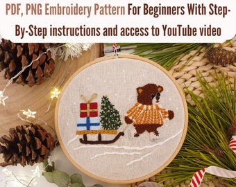 Könnte beinhalten: Stickrahmen-Kunst mit Stickerei, die einen braunen Bären zeigt, der einen Pullover trägt und einen Schlitten mit Geschenken und einem Weihnachtsbaum zieht. Der Rahmen befindet sich auf einer Holzoberfläche mit Kiefernästen und einem rot-weißem Band.