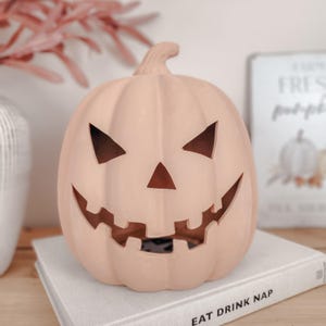 May include: A matte light peach-coloured ceramic jack-o'-lantern with a carved triangular nose, two triangular eyes, and a wide, jagged toothy grin. The pumpkin features vertical ridges and a small stem at the top, resting on a white book.