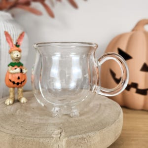 May include: A clear, double-walled glass mug shaped like a small cauldron with a rounded body, a wide rim, and a curved handle. The mug features three small, rounded feet at the base, providing stability while maintaining a smooth, transparent aesthetic.