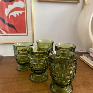 May include: Six olive green vintage drinking glasses with a textured, geometric pattern. The glasses are on a wooden surface, with framed artwork in the background. The glasses have a pedestal base and a wide rim.