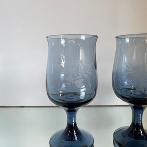 May include: Two blue-tinted stemmed glasses with etched floral designs. The glasses have a rounded bowl shape and a dark blue stem and base. The etched design is visible on the bowl of the glass. The glasses are likely for serving beverages.