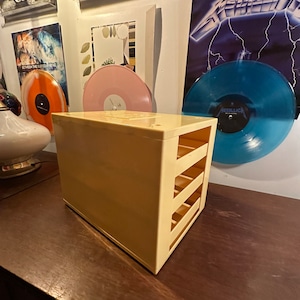 May include: A light yellow vinyl record storage unit with open slots on the front. The unit is rectangular and sits on a dark wooden surface. Vinyl records and framed art are visible in the background.