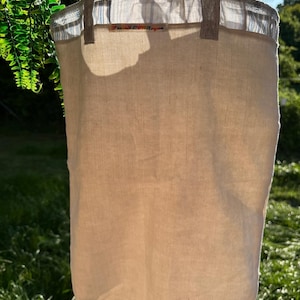 Könnte beinhalten: Eine beigefarbene Leinen-Tragetasche mit stabilen Griffen. Der obere Rand hat ein gestreiftes Stoffdetail und ein kleines Etikett. Die Tasche hängt im Freien, mit grünem Laub im Hintergrund.