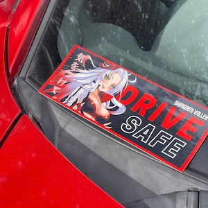 Puede incluir: Una pegatina negra y roja con el texto "DRIVE SAFE" y las palabras "SHINGAMI VALLEY" en blanco. La pegatina presenta una ilustración de estilo anime de una mujer con cabello largo y blanco y ojos verdes.