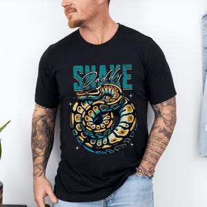 Op de afbeelding: Zwart T-shirt met een afbeelding van een opgerolde slang met blauwe, gele en bruine details. De tekst "Snake Daddy" staat boven de slang in teal en wit. Het T-shirt heeft een ronde hals.