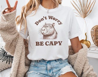 May include: White t-shirt with the text "Don't Worry BE CAPY" in brown lettering. The shirt features a smiling capybara illustration. The model is wearing blue denim shorts and a beige cardigan.