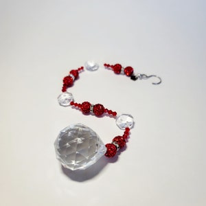 May include: A crystal suncatcher with a large, faceted teardrop-shaped crystal hanging from a string of red and clear beads. The beads are strung on a silver wire with a silver hook at the top.
