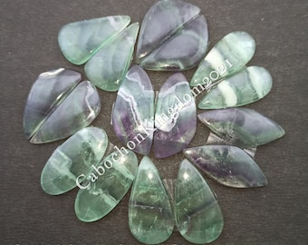 Può includere: Una collezione di 10 cabochon di fluorite verde, viola e bianco, ciascuno tagliato a forma di lacrima. I cabochon sono disposti in un cerchio su uno sfondo scuro.