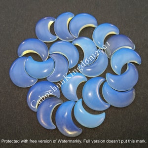 May include: A collection of blue crescent moon cabochons in varying shades of blue, arranged on a dark surface. The cabochons have a smooth, polished appearance, with a lighter, almost yellow, inner curve. The image includes the text "CabochonKingdom2021".