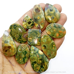 May include: A collection of ten green, brown, and yellow gemstone cabochons with a variety of shapes and sizes. The cabochons are displayed on a hand.