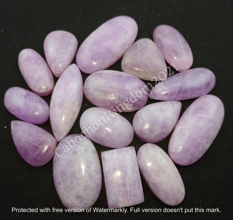 May include: A collection of polished, lavender-colored gemstones in various shapes, including ovals, teardrops, and rectangles. The stones have a smooth, glossy surface and are arranged on a dark background. The text "CabochonKingdom2021" is visible.
