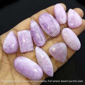 May include: A collection of polished, light purple gemstones in various shapes, including oval, teardrop, and rectangular. The stones are held in a hand, with the text "Cabochon Kingdom2021" visible. The image also includes the text "Protected with free version of Watermarkly. Full version doesn't put this mark."