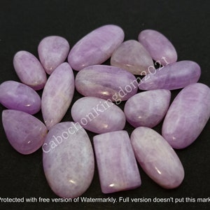 May include: A collection of polished, lavender-colored gemstones in various shapes, including ovals, teardrops, and rectangles. The stones have a smooth, glossy surface and are arranged on a dark background. The image includes the text "CabochonKingdom2021".