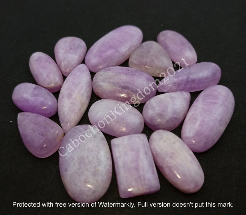 May include: A collection of polished, lavender-colored gemstones in various shapes, including ovals, teardrops, and rectangles. The stones have a smooth, glossy surface and are arranged on a dark background. The image includes the text "CabochonKingdom2021".