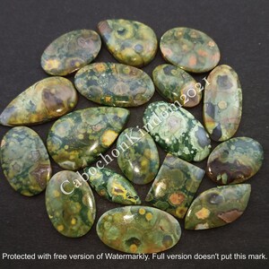 May include: A collection of 16 green, brown, and yellow polished gemstone cabochons. The cabochons are in various shapes, including oval, teardrop, and rectangular. The cabochons are arranged in a circle on a black background.