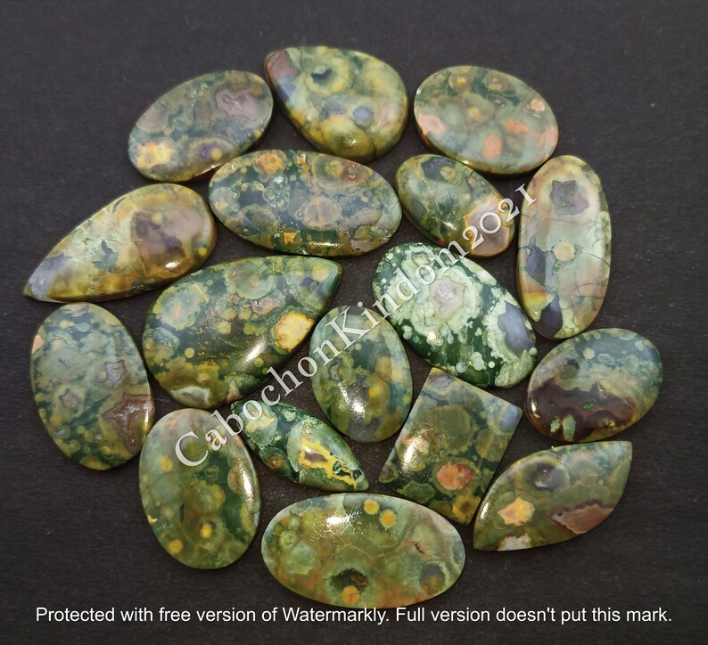 May include: A collection of 16 green, brown, and yellow polished gemstone cabochons. The cabochons are in various shapes, including oval, teardrop, and rectangular. The cabochons are arranged in a circle on a black background.