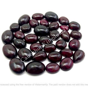Könnte beinhalten: Eine Sammlung loser, polierter, dunkelroter Granat-Cabochons in verschiedenen ovalen und tropfenförmigen Formen. Die Steine haben eine glatte, glänzende Oberfläche und tiefe burgunderrote Töne. Der Text CabochonKingdom21 ist über die Mitte gelegt.