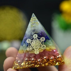 May include: A small, multi-colored crystal pyramid with a gold geometric design in the center. The pyramid is made of clear resin and has a layer of gold glitter around the base.