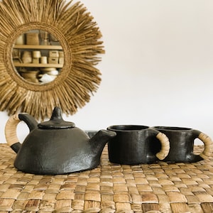 May include: A black ceramic teapot and two mugs with woven handles sit on a woven straw mat. The teapot has a rounded body and a spout on the side. The mugs have a simple cylindrical shape.