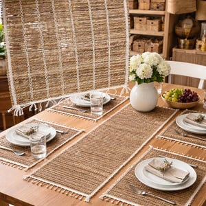 Op de afbeelding: Een eettafelset met geweven placemats, een tafelloper en een bijpassend hangstuk. De tafel is gedekt met witte borden, bestek en glazen. Een witte vaas bevat witte bloemen en een schaal met druiven.