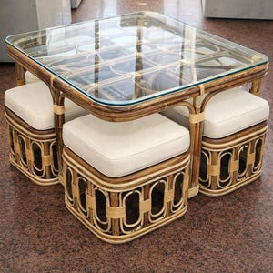 May include: A square coffee table with a glass top and a dark brown rattan frame, featuring four matching square stools with off-white upholstered cushions that tuck neatly underneath the table frame for compact storage.