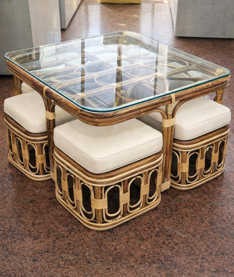 May include: A square coffee table with a glass top and a dark brown rattan frame, featuring four matching square stools with off-white upholstered cushions that tuck neatly underneath the table frame for compact storage.