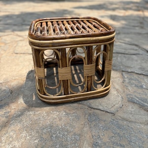 May include: A square stool constructed from dark brown and light tan rattan. The top features a slatted surface, while the sides are composed of vertical oval-shaped frames reinforced with horizontal rattan bindings at the center and corners.