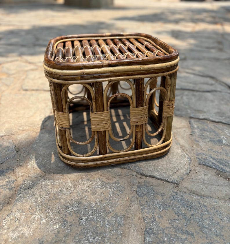 May include: A square stool constructed from dark brown and light tan rattan. The top features a slatted surface, while the sides are composed of vertical oval-shaped frames reinforced with horizontal rattan bindings at the center and corners.